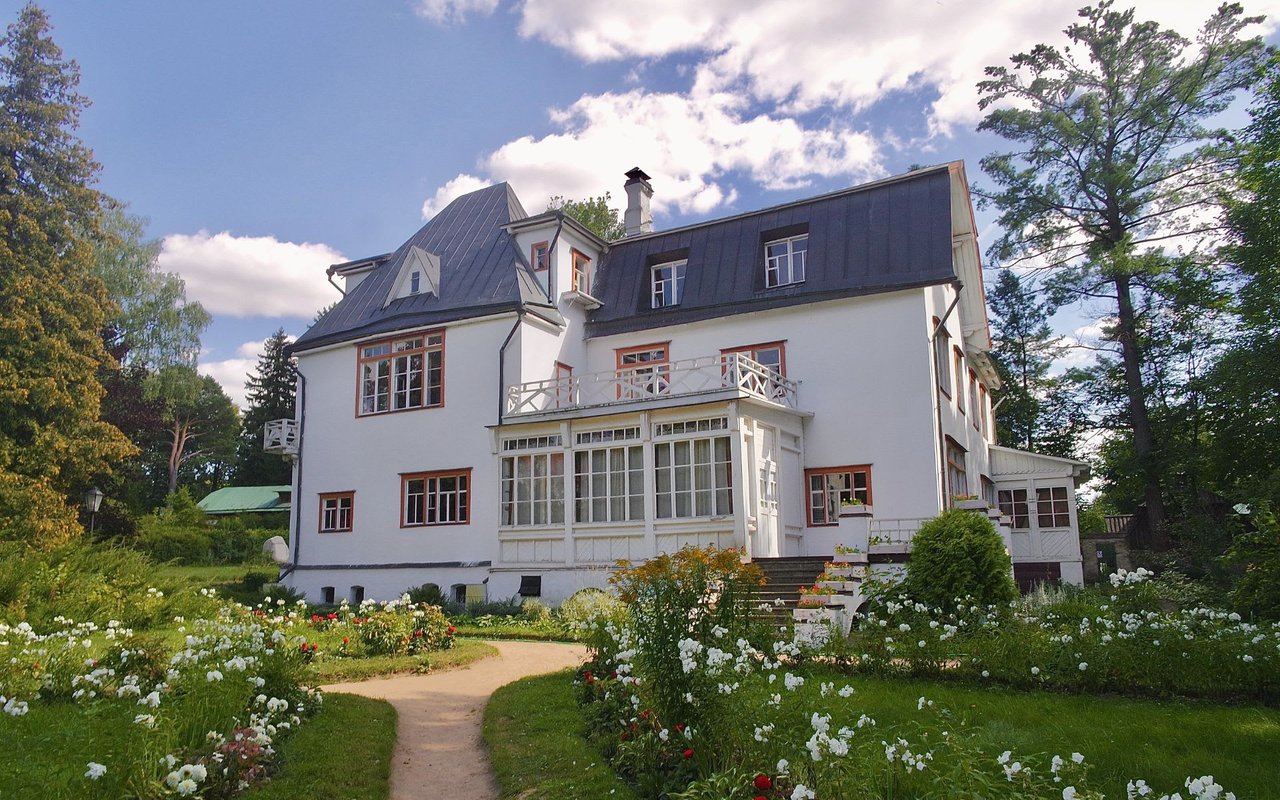  A grand, white multi-story house with a dark gray roof stands amidst a vibrant green garden under a bright, partly cloudy sky. 