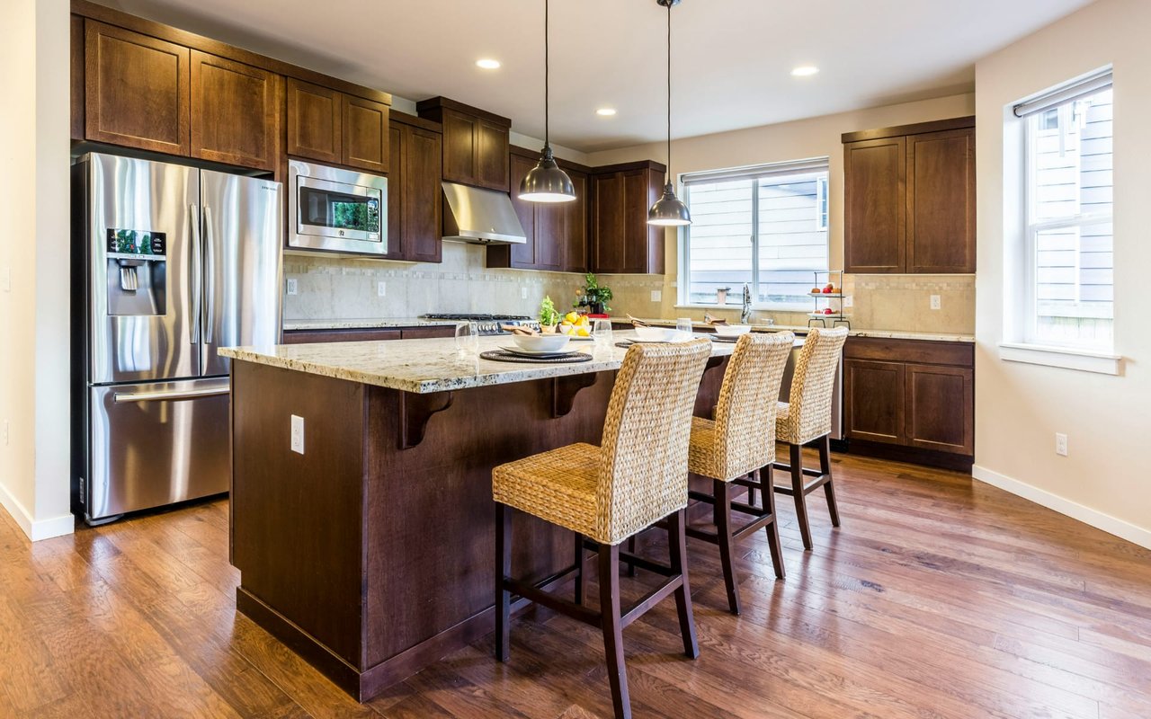 Kitchen with a large granite island, dark wood cabinetry, stainless steel appliances, and woven bar stools.