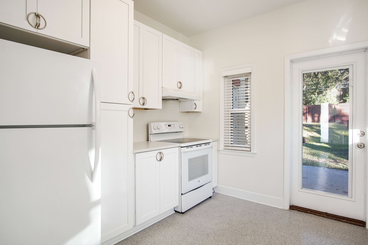 A kitchen area with white cabinetry and appliances featuring a stove, refrigerator, and cabinets. The room has a door leading outside, providing access and natural light.