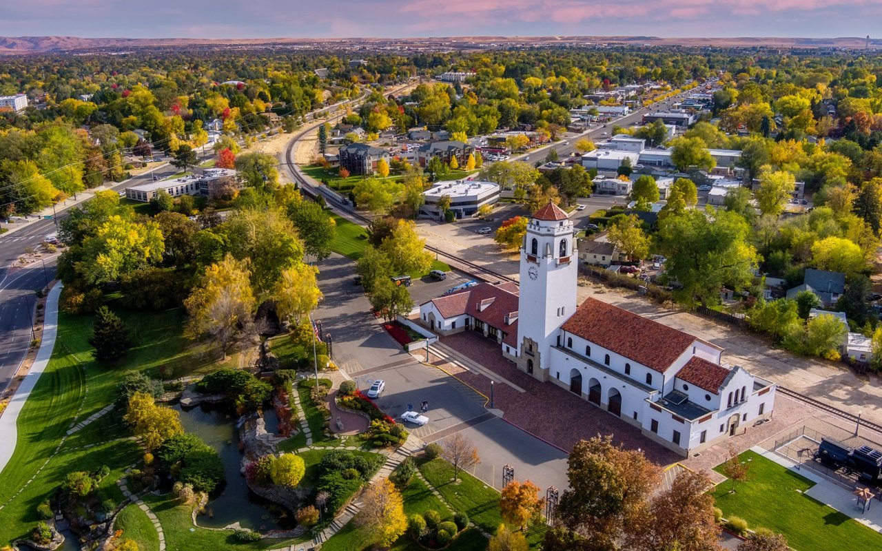  An aerial view of a historic train depot with a white stucco building and a prominent clock tower topped with a red tile roof.