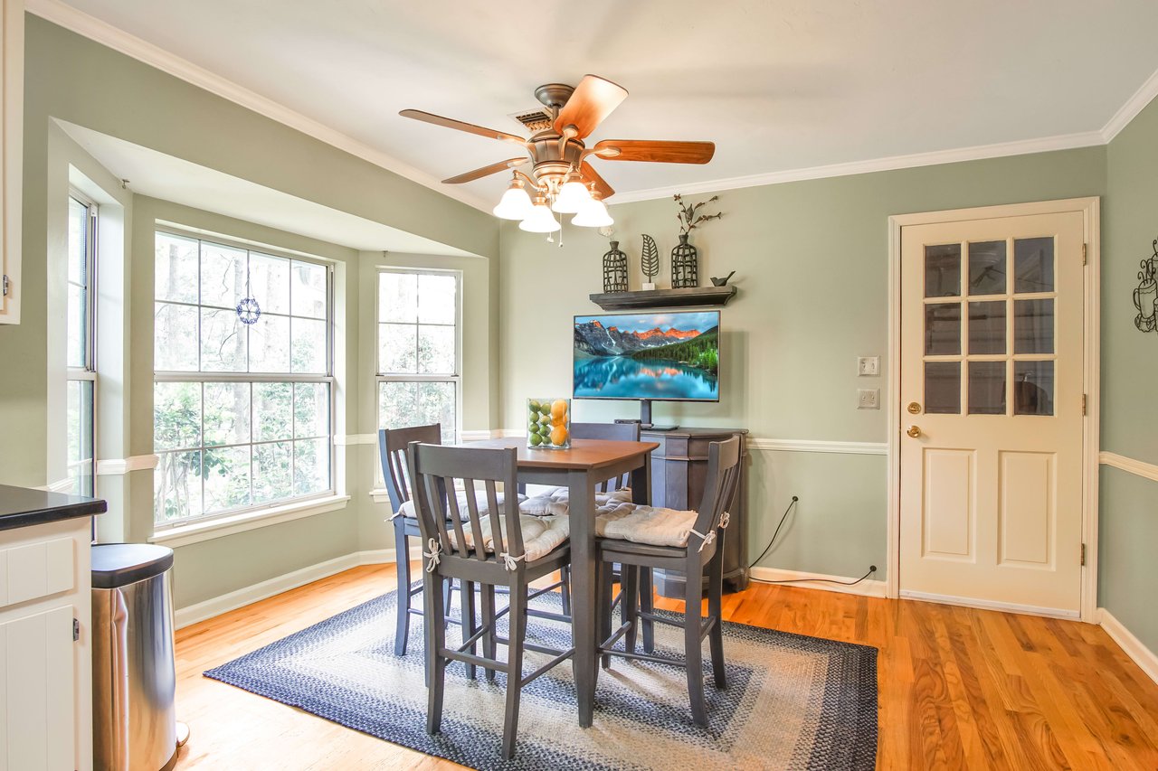 An interior shot of a dining area with a table and chairs. The room has large windows letting in natural light and a ceiling fan. There is a door leading outside.