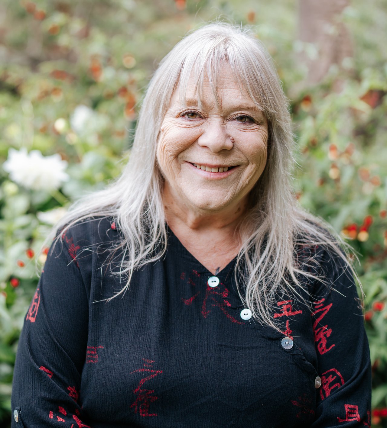 Joyce Rinehart smiles in a black top with red designs, photographed against a garden backdrop.