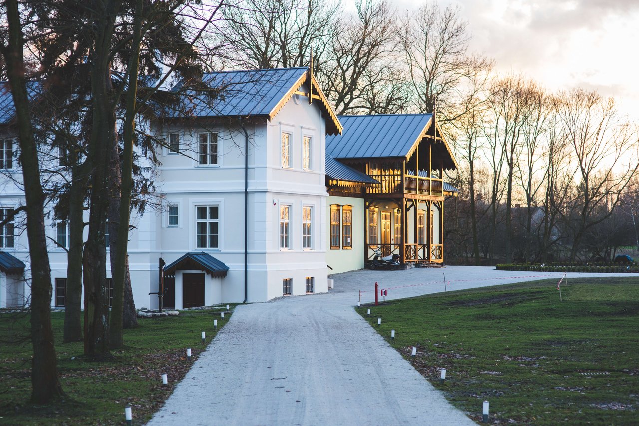 A white house with a gray roof stands beside a wooden house with a balcony, surrounded by a gravel driveway and green lawns.