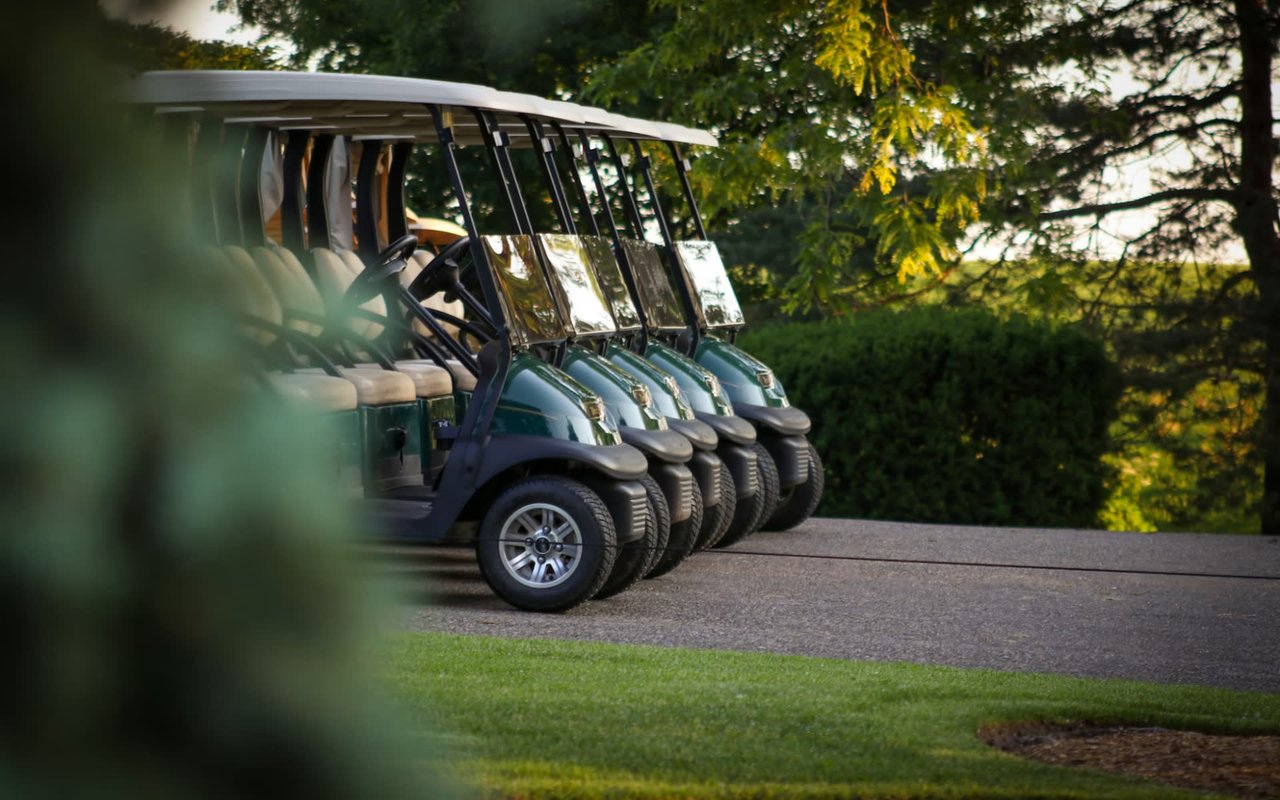  A line of five dark green golf carts with white roofs are parked in a row on an asphalt path next to a well-maintained green lawn.