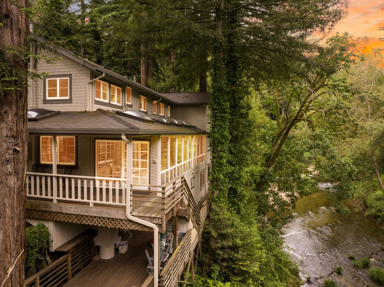 Wooded home with wraparound decks beside a creek at sunset.