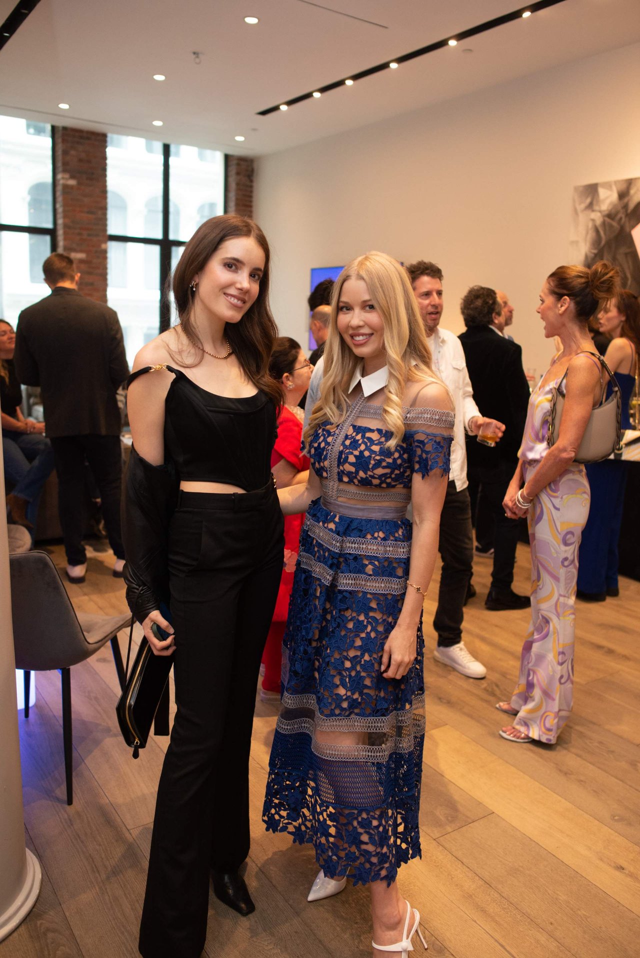Two women pose together at an indoor event, surrounded by mingling guests.