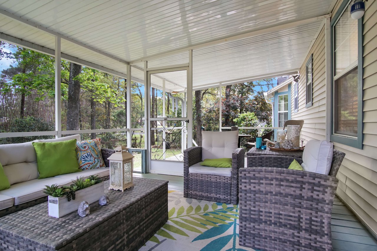 A screened porch or sunroom with wicker furniture and colorful cushions. The area is cozy and provides a pleasant view of the surrounding greenery.