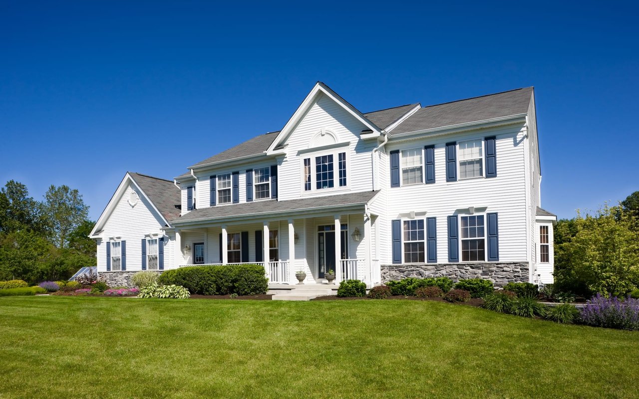 A two-story white house with blue shutters on a lush green lawn surrounded by various trees.