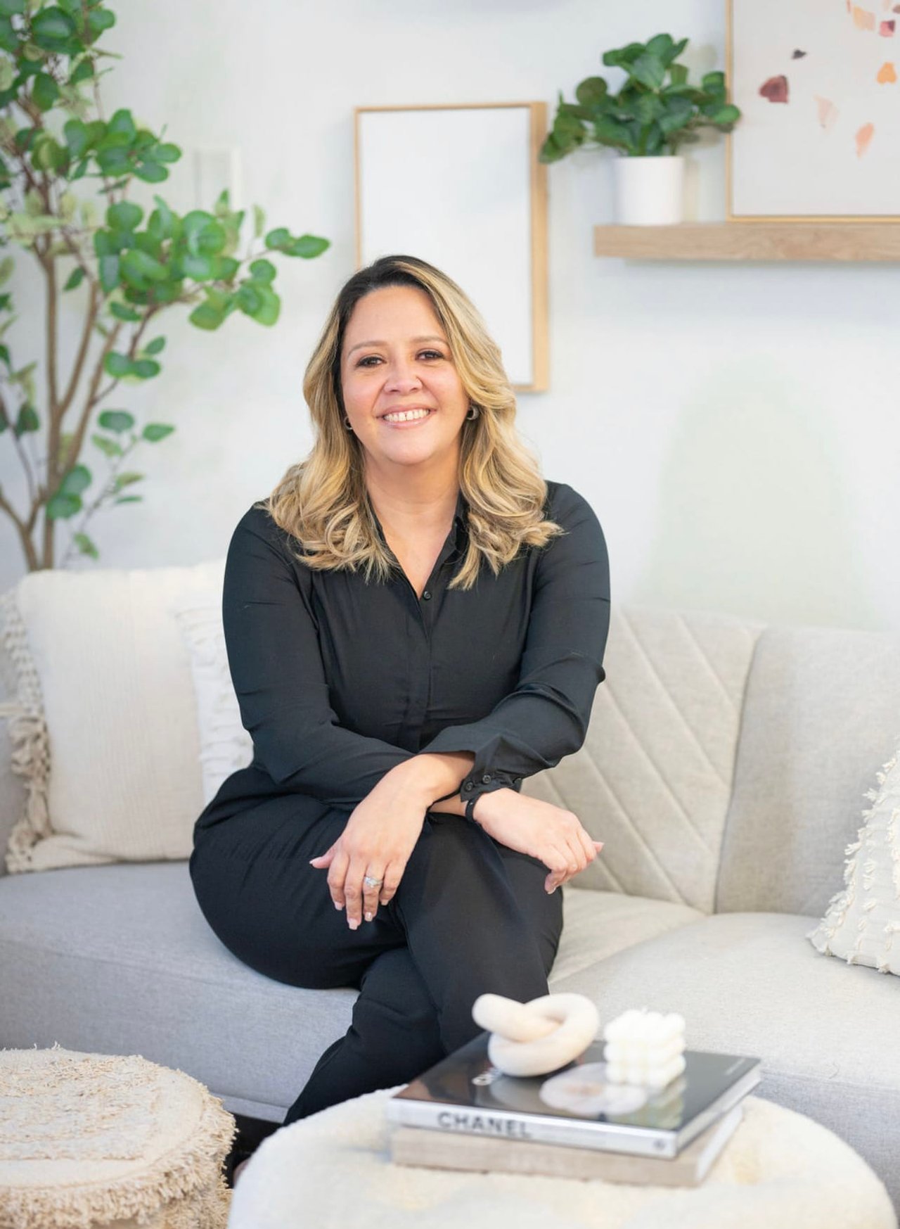 Smiling woman in black seated cross-legged on a light sofa beside a leafy plant.