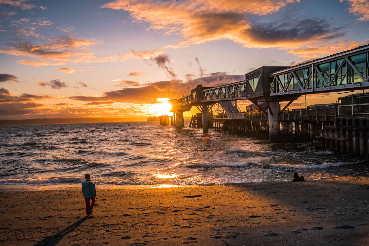 A young child walks along a sandy beach towards a pier at sunset