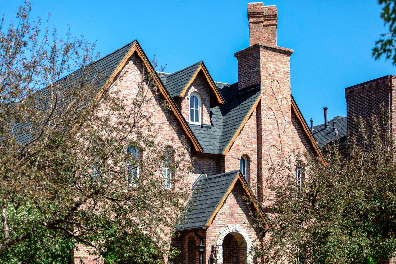 A large brick house with a black roof and chimney, surrounded by trees on a sunny day.