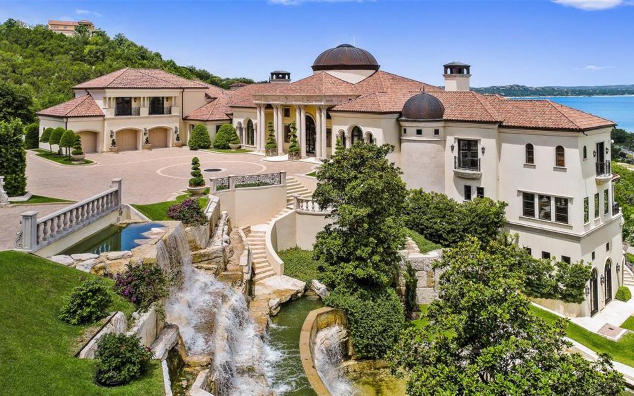 A grand Mediterranean-style mansion with a circular driveway, cascading waterfalls, and stunning lake views.