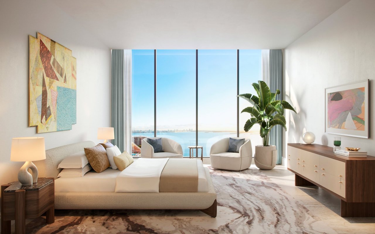 Staged bedroom with a king bed and lounge chairs overlooking the water through floor-to-ceiling windows.