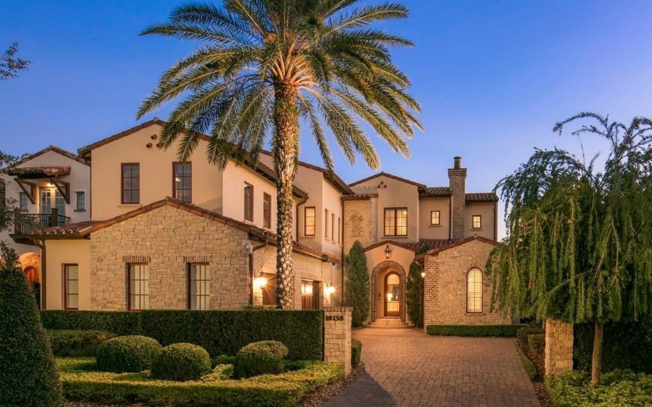 A large, two-story white house with a red tile roof, a driveway, a curved sidewalk, and a large palm tree in front.