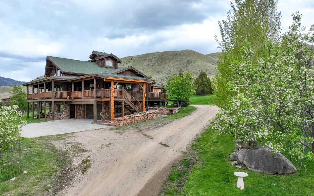  A multi-story wooden house with a dark green roof and a large wraparound porch supported by wooden pillars. 