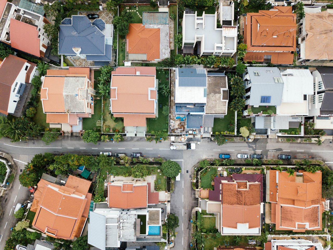 Aerial view of closely spaced homes with colorful roofs, leafy yards, and parked cars along the street.