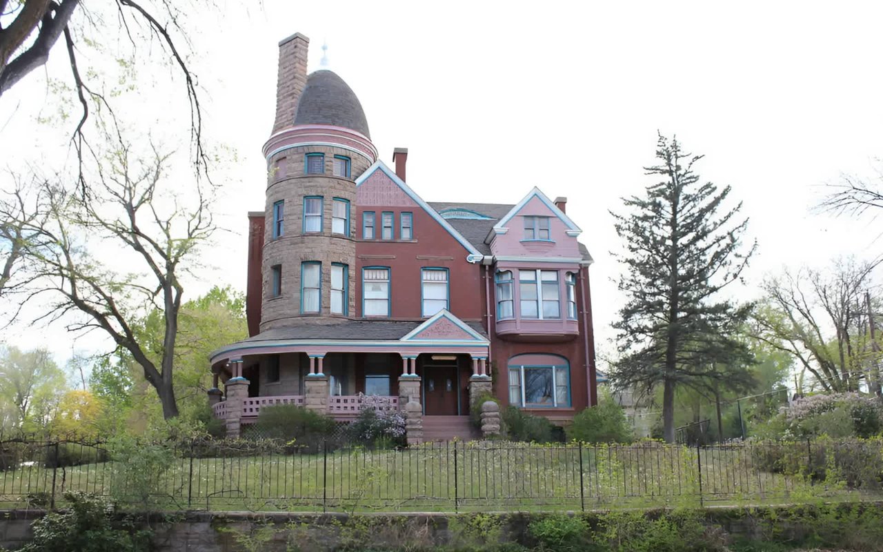 Historic Victorian-style mansion with a round turret, arched windows, and a wrap-around porch, surrounded by mature trees.