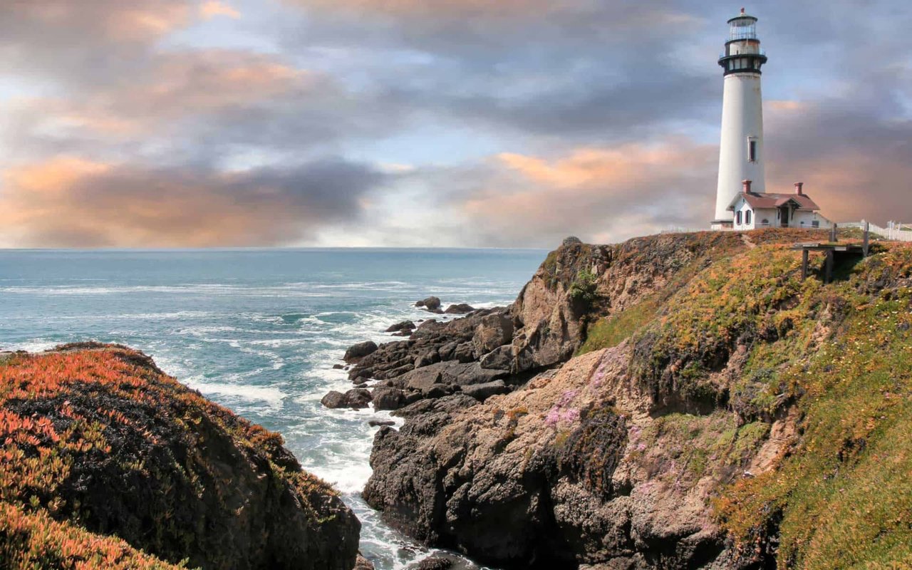 Pigeon Point Lighthouse perched on coastal cliffs overlooking the Pacific Ocean in Pescadero, California, with waves crashing below and a dramatic sky above.