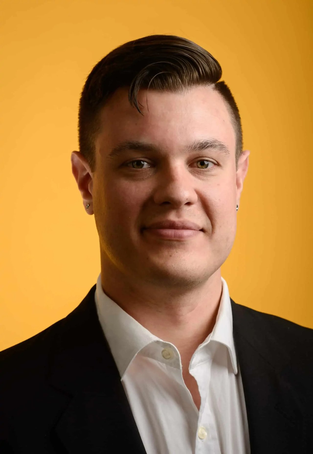 A formal photo of Kelly Chavers dressed in a formal suit with a white dress shirt underneath against a solid, vibrant yellow background. 