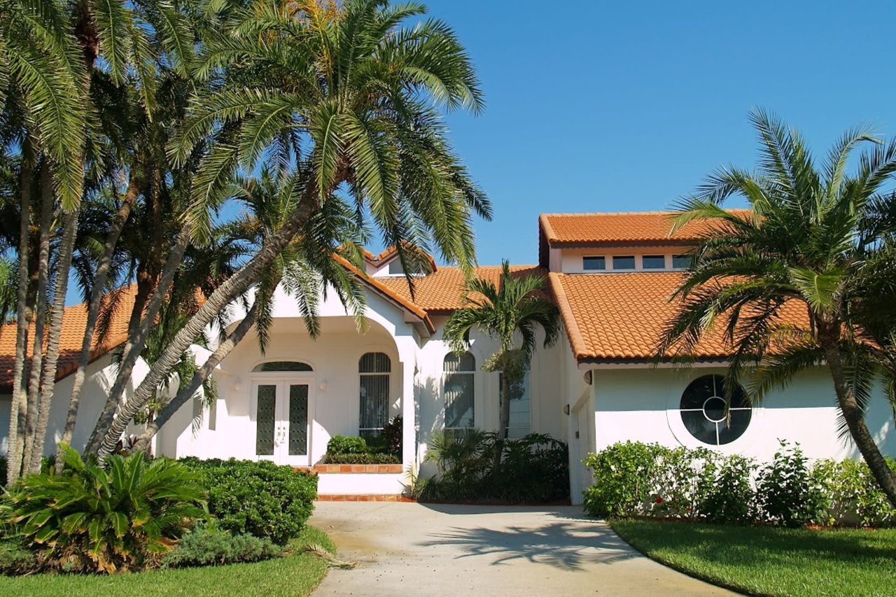 A white house with a red tile roof surrounded by palm trees. The house has a driveway and a lush green lawn.