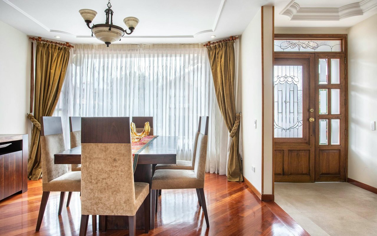 A formal dining room with wooden floors, a dining table and chairs, a hanging chandelier, and a large door to the left.
