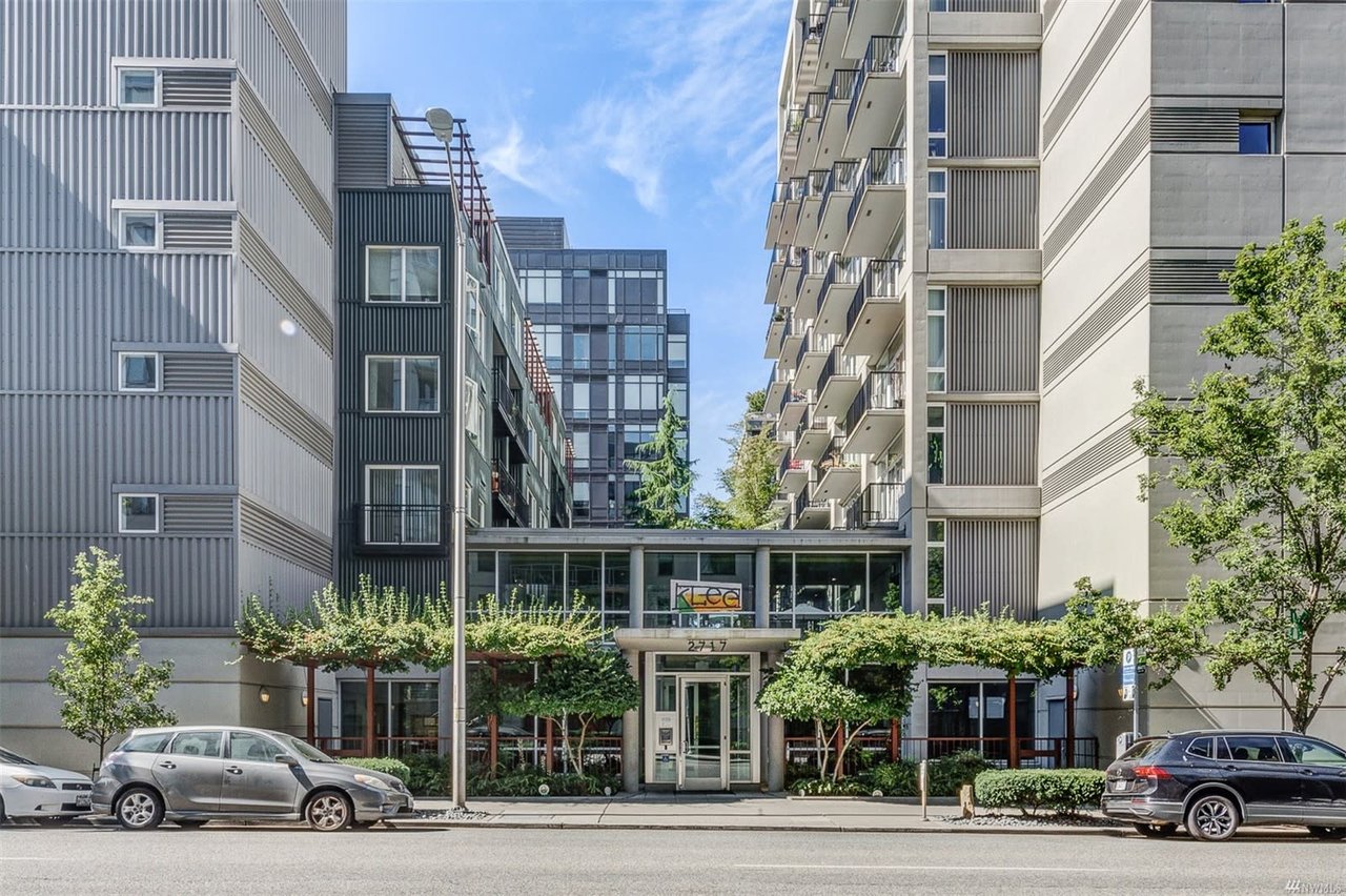 Modern condo entrance with a glass canopy flanked by industrial-style buildings and lush greenery.