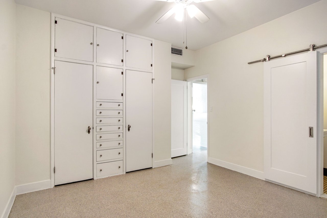 An interior shot of a bedroom with built-in storage and a sliding barn door leading to a bathroom. The room has a ceiling fan and a neutral color scheme.