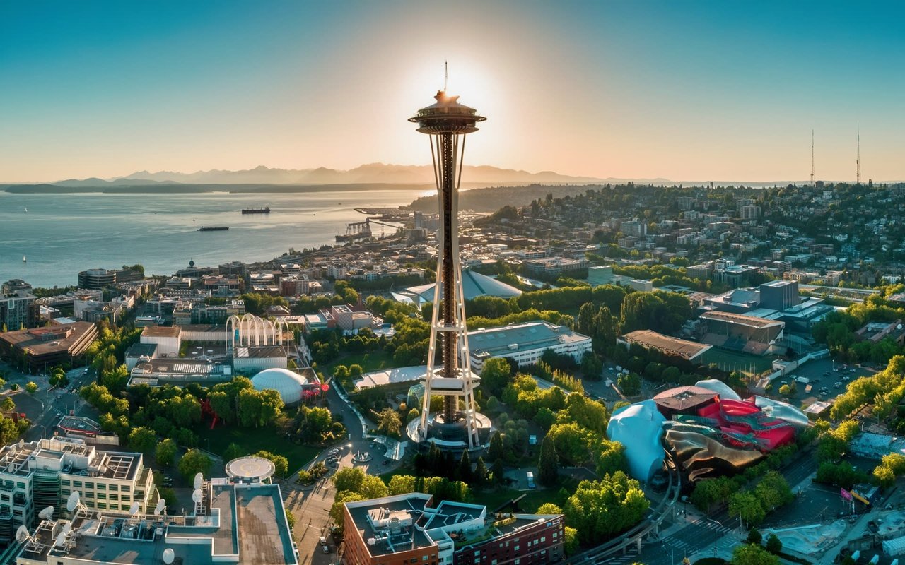  An aerial view of the Seattle skyline with the Space Needle prominently centered, its top observation deck silhouetted against the setting or rising sun. 