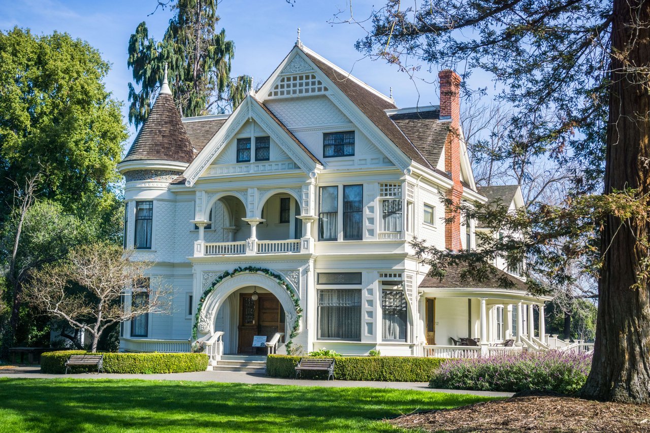 A large Victorian house with a tower, surrounded by lush greenery in a park-like setting.