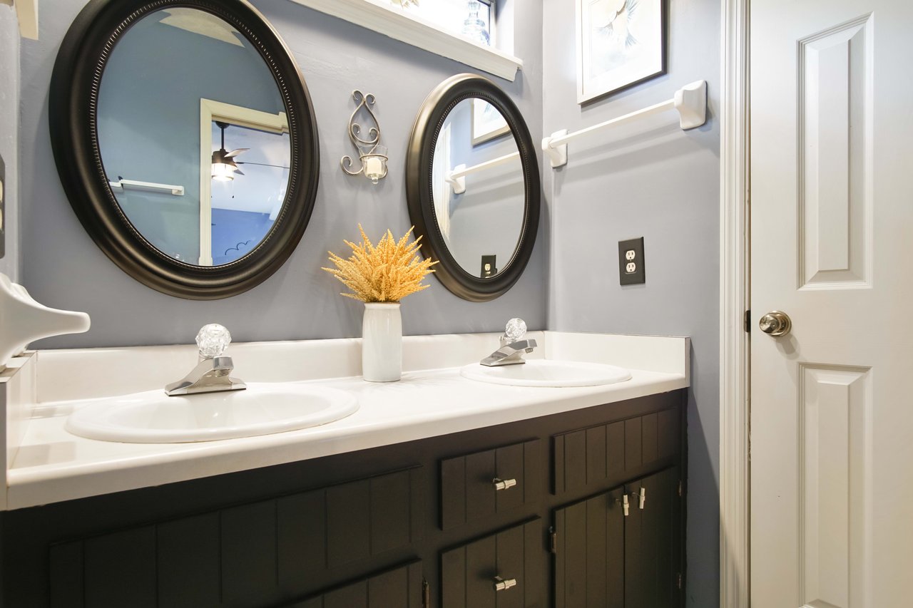 A bathroom with double sinks, each with a round mirror. The bathroom has dark cabinets, white countertops, and modern fixtures.