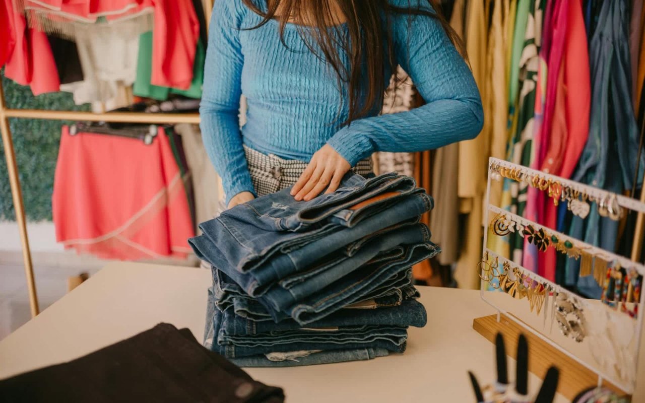  A person wearing a blue long-sleeved shirt and checkered pants is standing behind a white table, folding or arranging a large stack of blue jeans. 