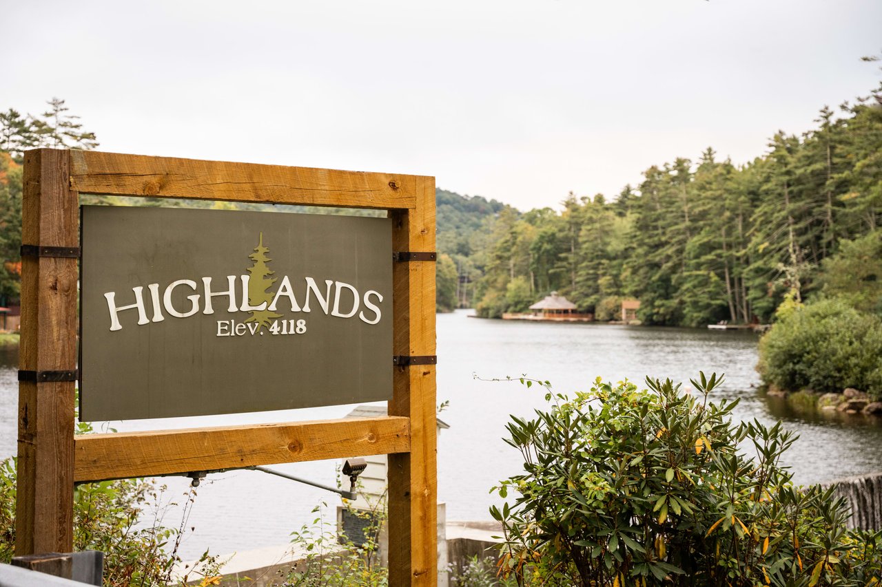 Wooden sign 'Highlands Elev: 4118' before a serene lake, trees, and charming house—ideal for real estate or serene work environment ads.