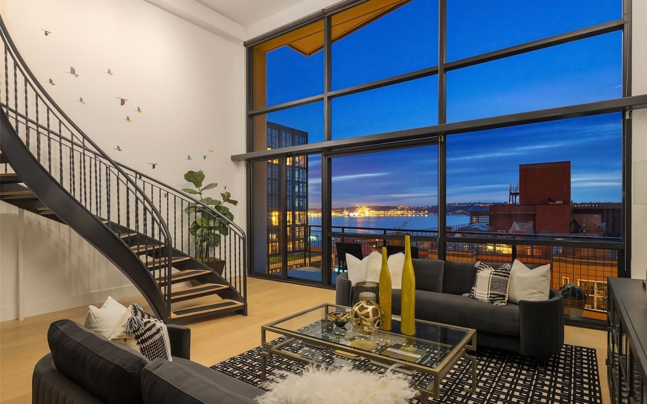  An expansive living room features a sweeping view of a city waterfront at dusk through large, multi-paned windows.