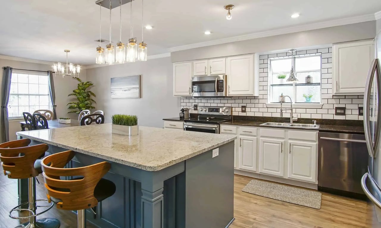 A modern kitchen with a large island in the center. The kitchen features white cabinets, stainless steel appliances, and bar stools around the island. The space is bright and well-lit.