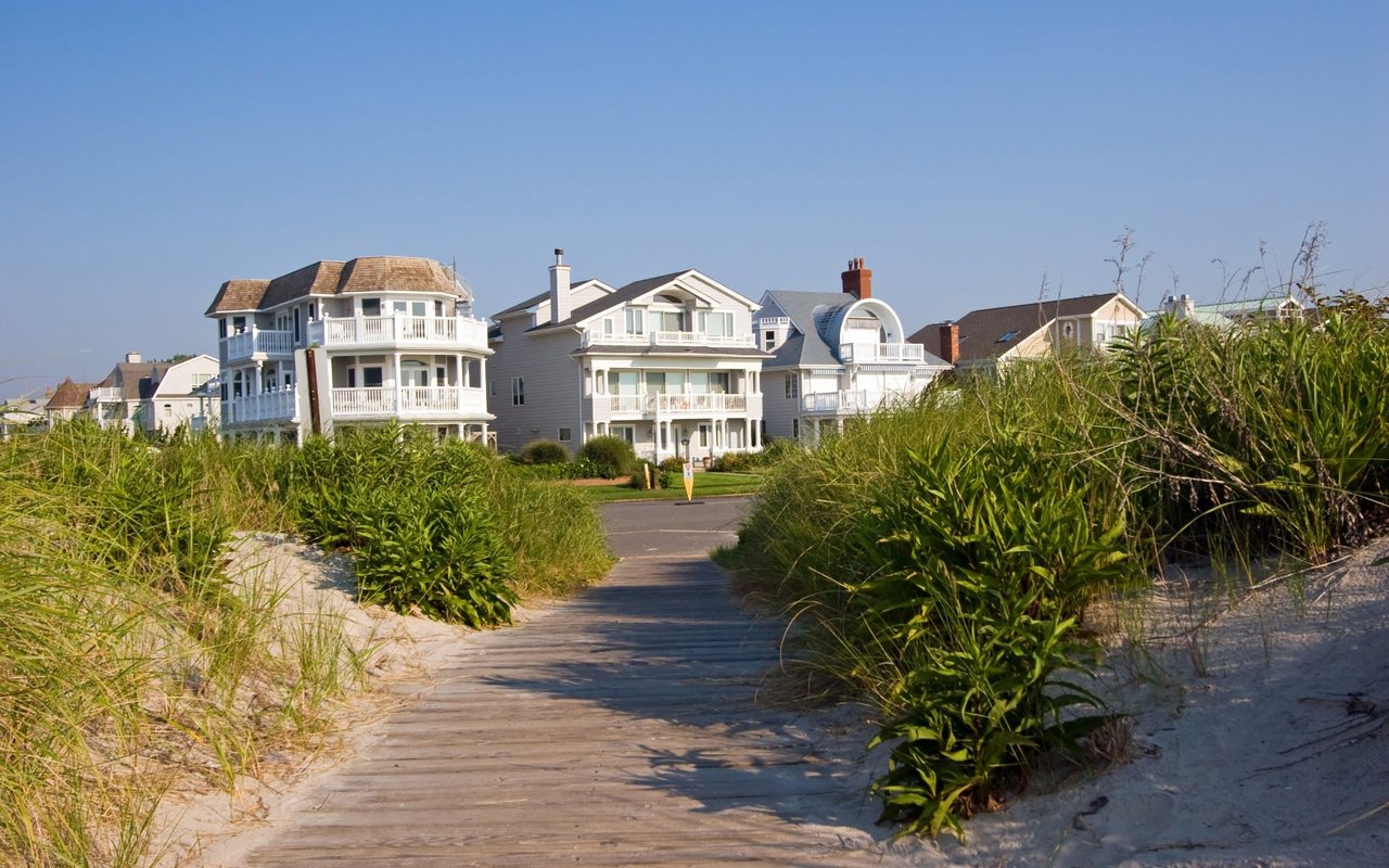 Coastal homes beyond a wooden boardwalk winding through sandy dunes and beach grass.