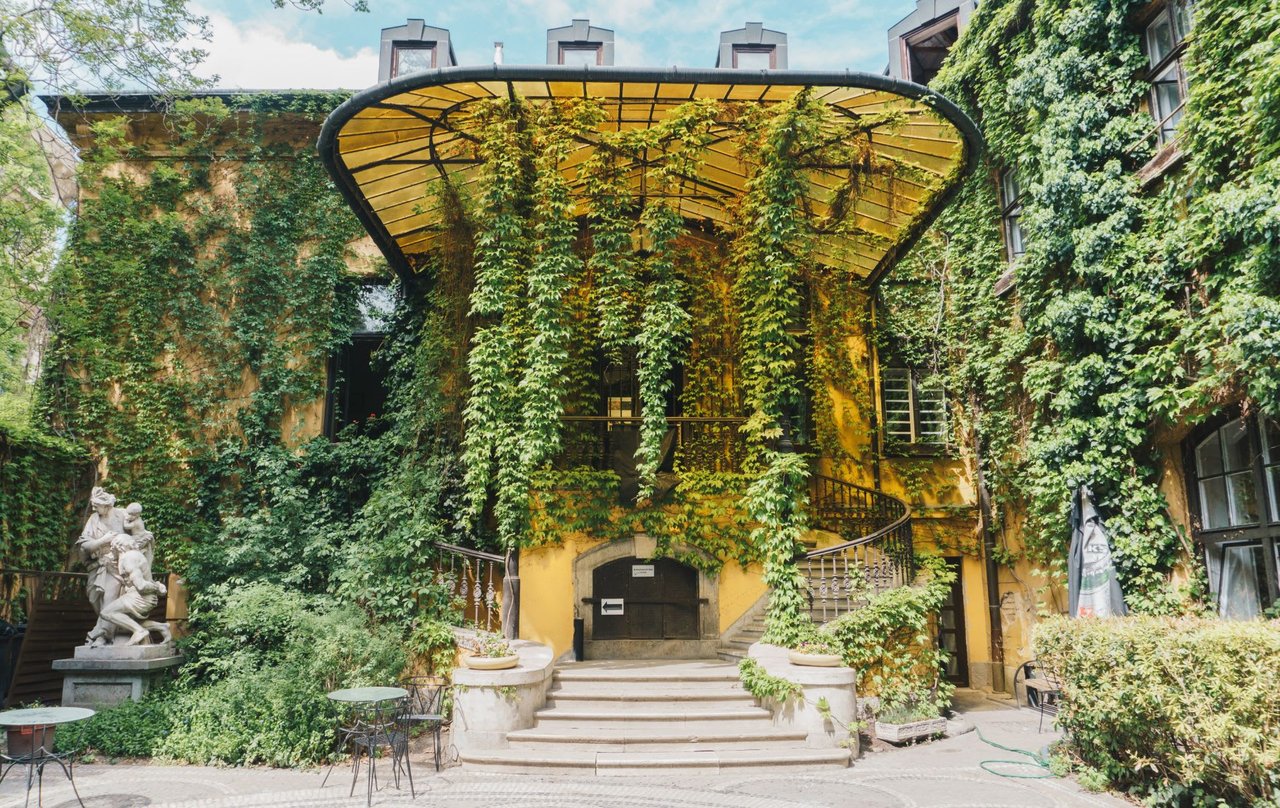  A grand, multi-story building with a warm yellow facade is almost entirely covered in lush green ivy, creating a vibrant, overgrown appearance. 