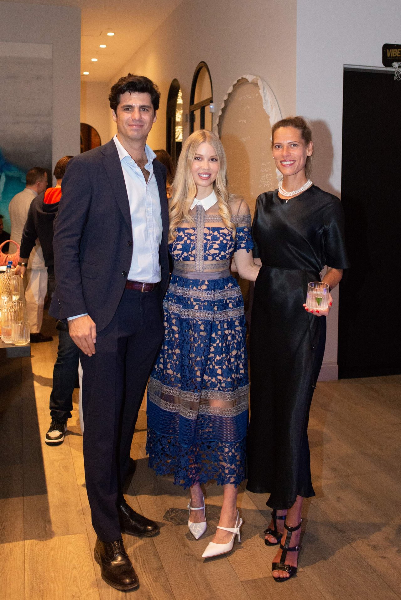 Three adults pose together at an indoor event; the women wear blue lace and black dresses.