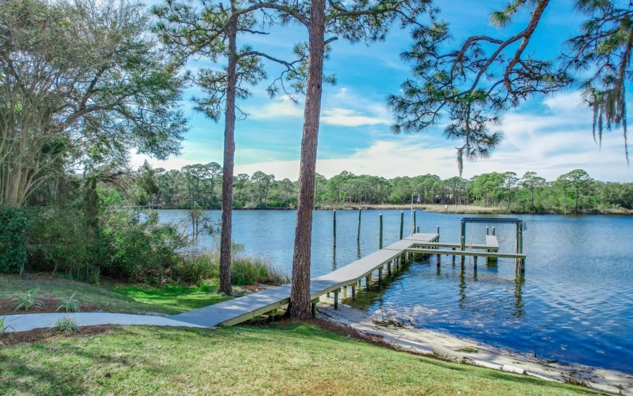 A wooden dock extends into a calm, blue lake surrounded by lush greenery and tall pine trees.