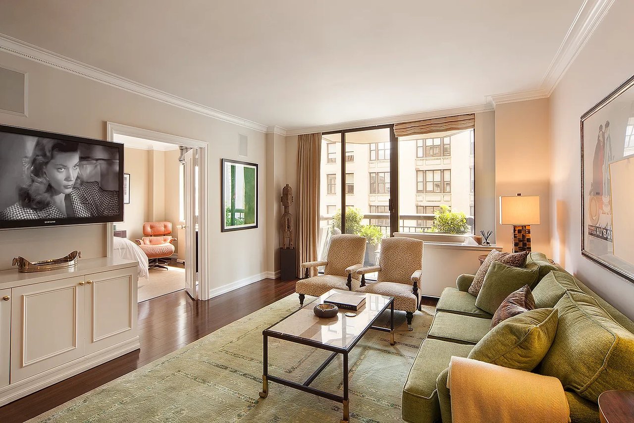 Living room with green sofa, two armchairs, glass coffee table, and large windows overlooking neighboring buildings.