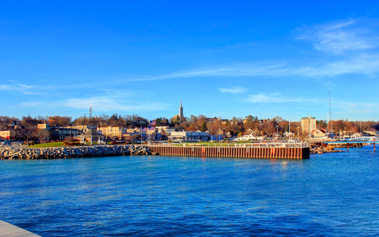 Coastal town with a clear blue sky, calm water, and buildings on a hill.