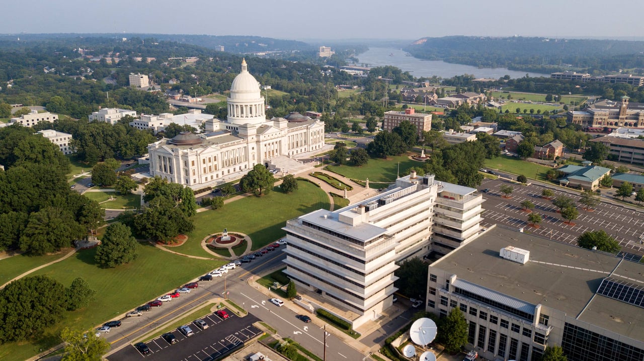 An aerial shot shows the Arkansas State Capitol building, a grand white neoclassical structure with a large dome