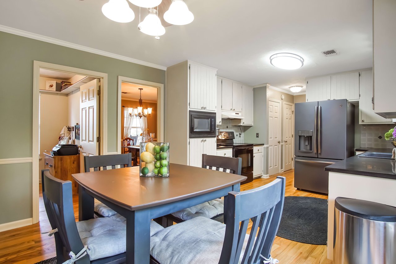 A dining area adjacent to a kitchen. The dining table has chairs around it, and the kitchen features white cabinets, stainless steel appliances, and a modern design.