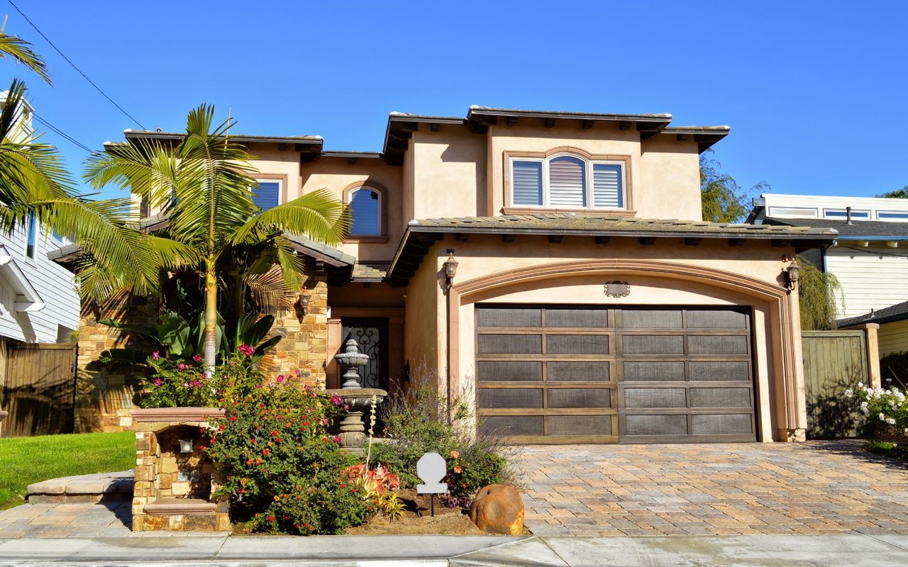 The front features a dark brown garage door with square panels and a stone-clad entrance area to the left, which includes a decorative fountain. 