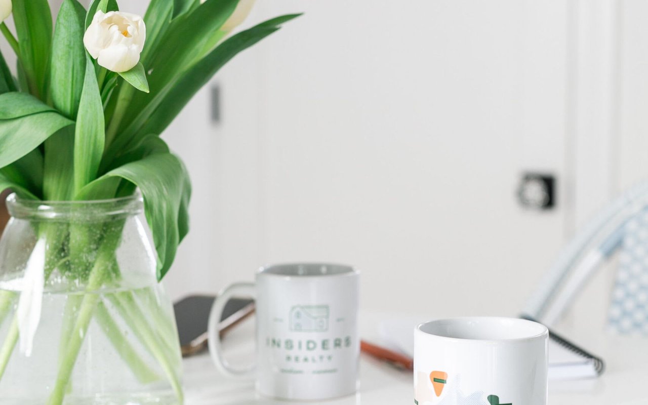 A vase filled with flowers sits on a table, alongside coffee mugs featuring the Insiders Realty logo.