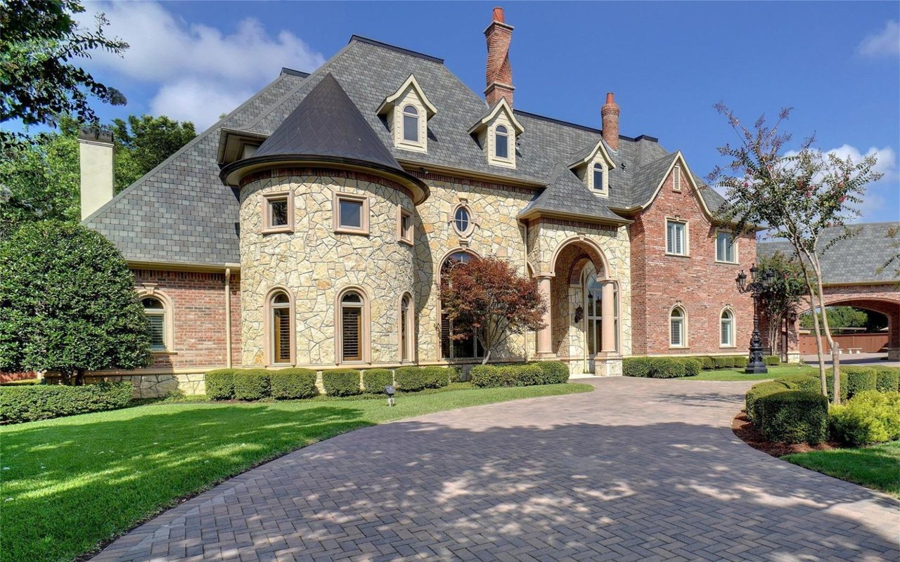A three-story Georgian mansion with a slate roof, chimneys, front porch, brick driveway, hedges, and trees in the front lawn.