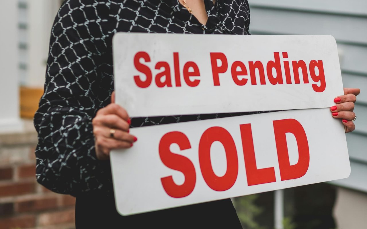 A woman holds a "Sale Pending" sign, indicating that a sale is in progress.