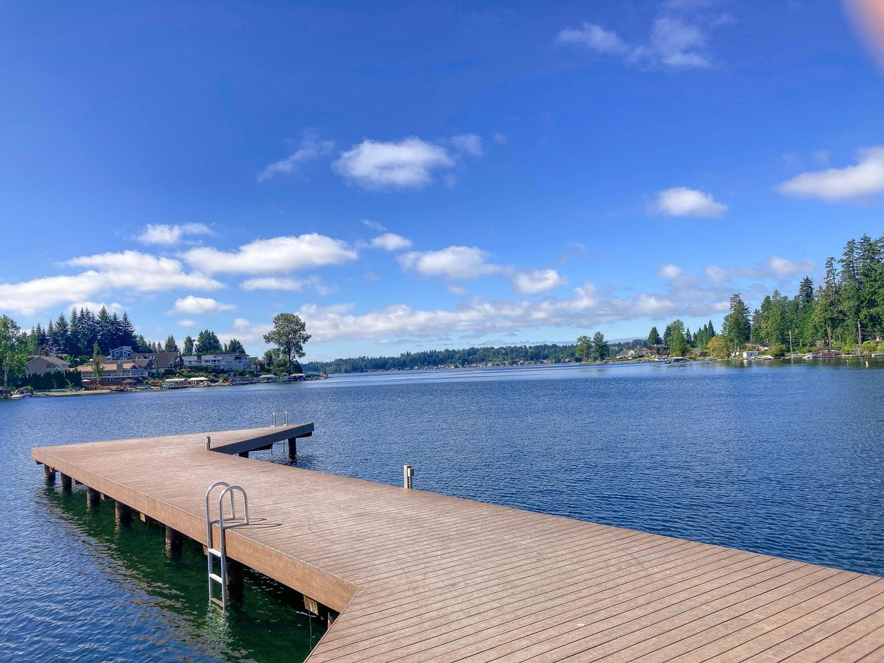 A wooden dock extends into a calm lake with a clear blue sky and fluffy white clouds