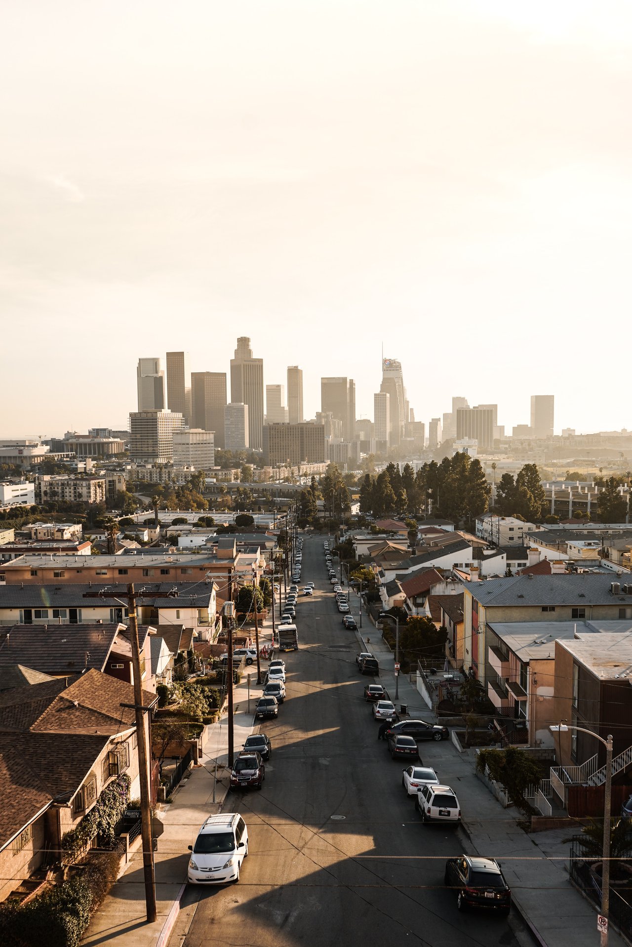 Downtown Los Angeles skyline beyond a residential street lined with houses and parked cars.
