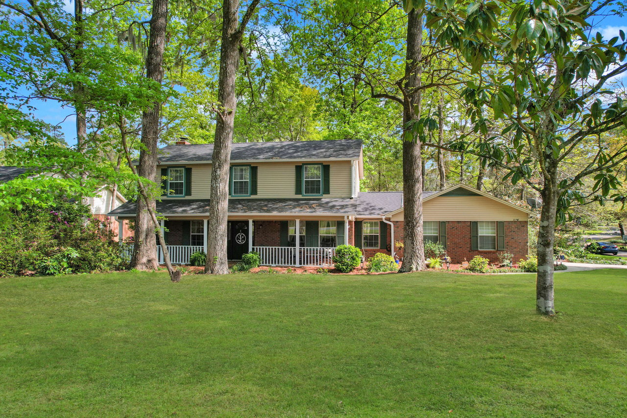 An exterior view of a large house with a well-maintained lawn. The house is surrounded by tall trees, providing a natural and serene setting.