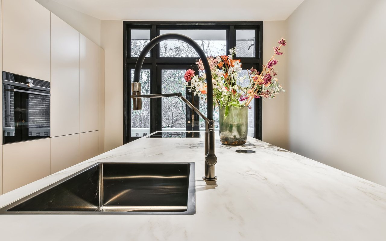 Modern kitchen with a cooktop, faucet, and a ceramic vase filled with flowers beside a large window offering an outdoor view.